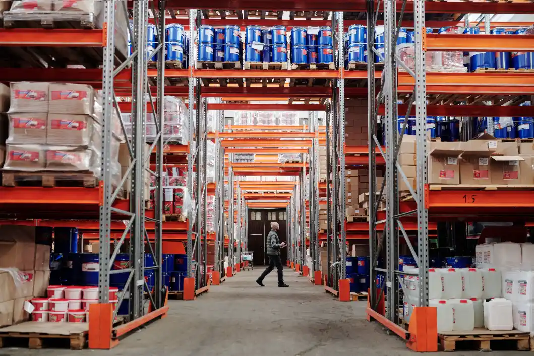 Corredor de um armazém com racks metálicos e prateleiras lotadas de caixas e materiais, com um funcionário em destaque ao fundo, indicando armazenagem terceirizada para empresas.