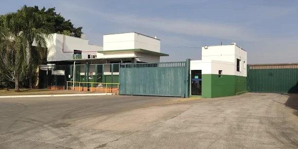 Entrada de uma unidade industrial moderna com portão verde e estrutura de metal, prédio branco e verde ao fundo, pátio de concreto e céu com nuvens.